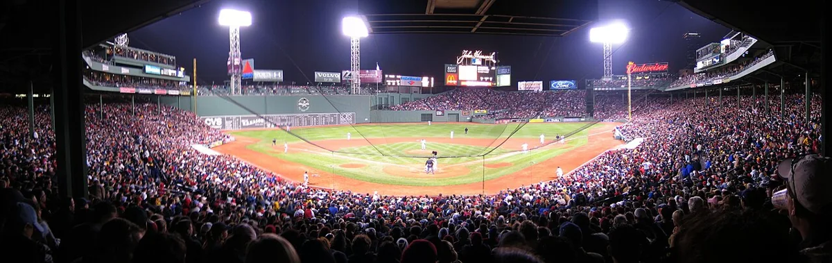 A packed Fenway Park under floodlights, with the baseball diamond in the center.