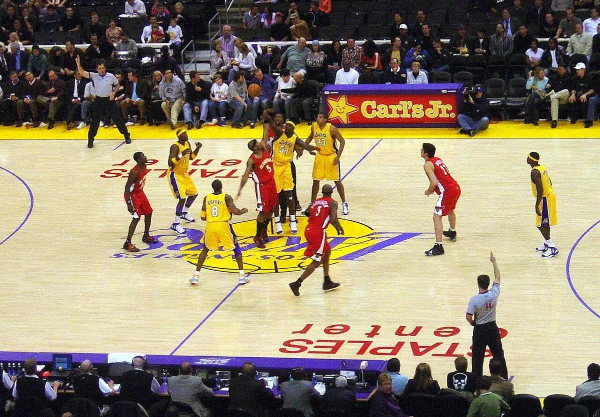 Lakers and Hawks players contest a jump ball at center court before a seated crowd.